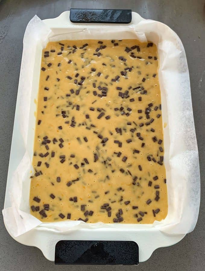 Orange Blondie with Dark Chocolate Chips A rectangular baking dish lined with parchment paper holds unbaked orange blondie batter with dark chocolate chips scattered throughout.