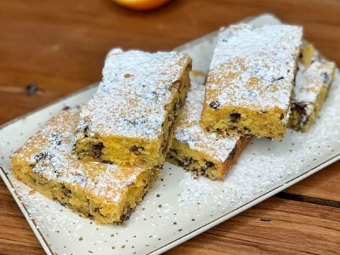 Rectangular orange blondie with dark chocolate chips, dusted with powdered sugar on a white plate, with a halved orange in the background.