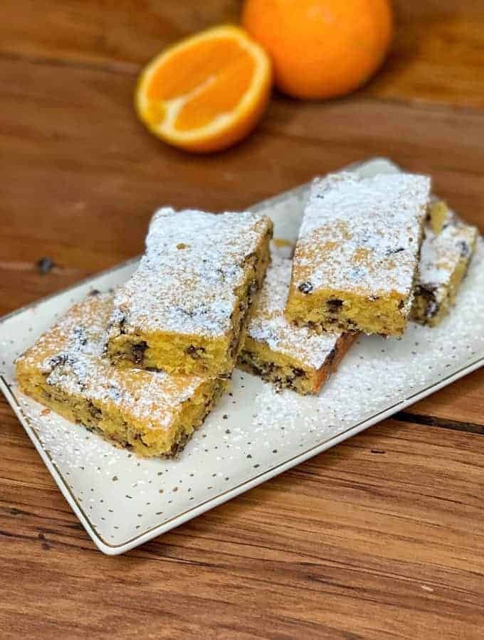 Rectangular orange blondie with dark chocolate chips, dusted with powdered sugar on a white plate, with a halved orange in the background.