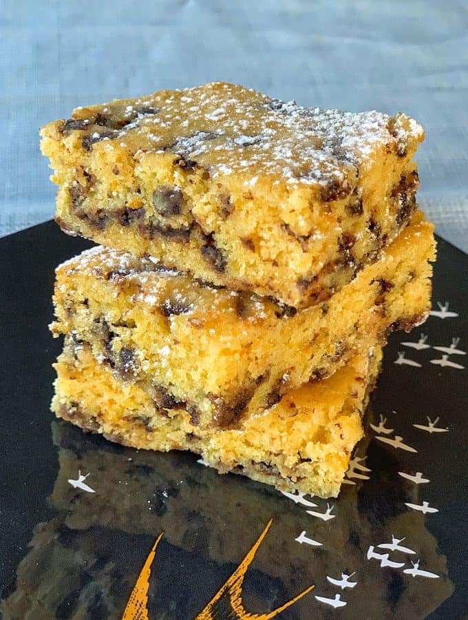 Orange Blondie with Dark Chocolate Chips Three orange blondie bars with dark chocolate chips stacked on a black plate with a bird design, dusted lightly with powdered sugar.