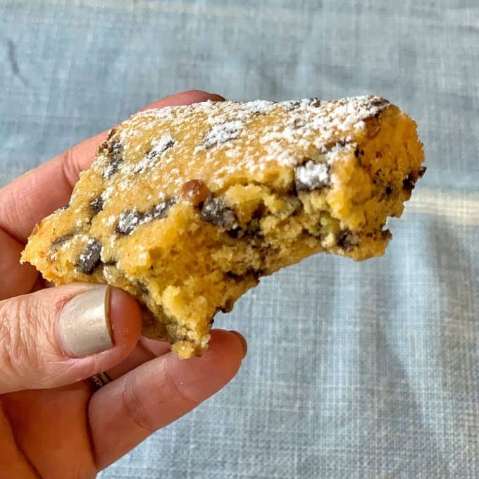 A hand holding a partially eaten orange blondie with dark chocolate chips, dusted with powdered sugar, against a light blue textured background.