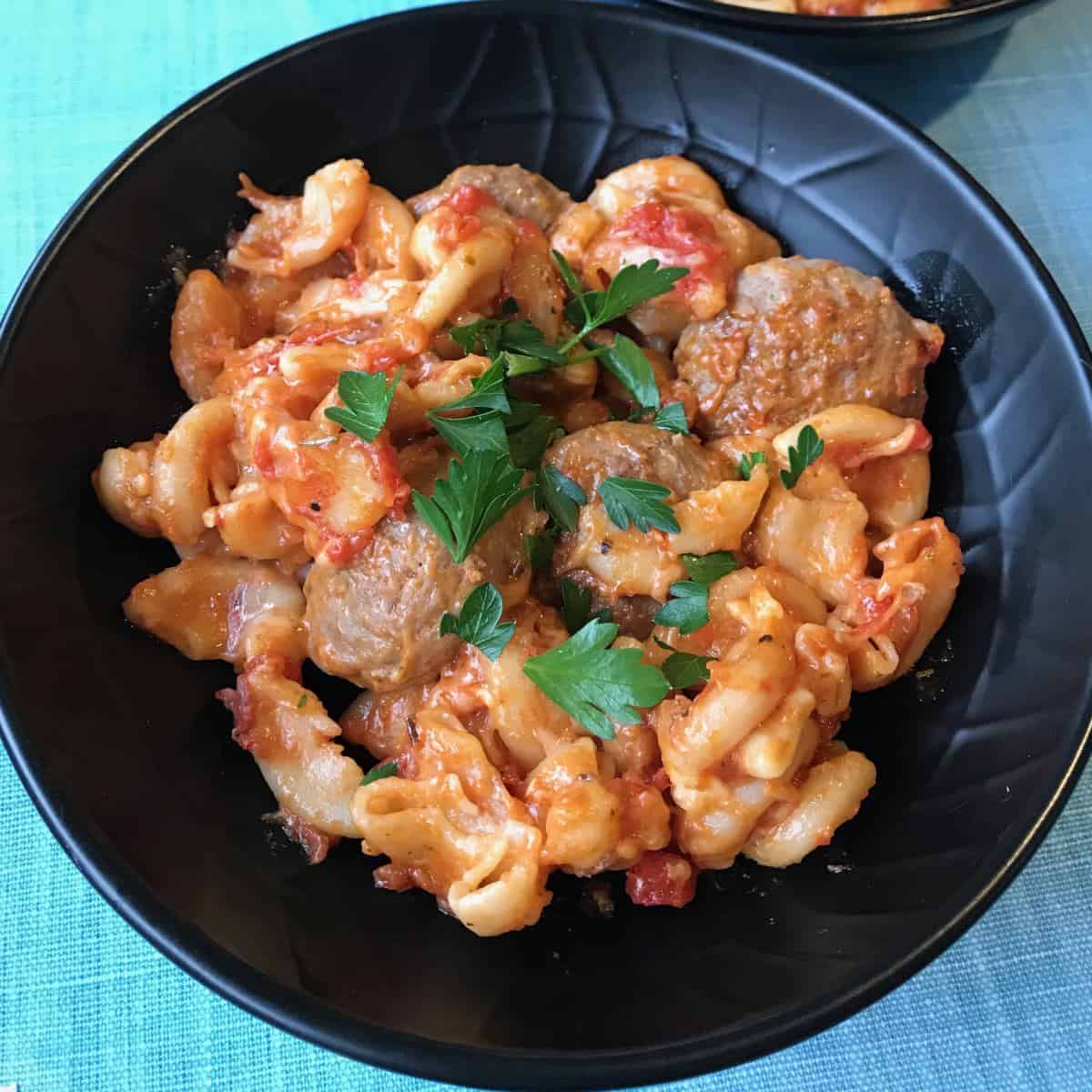 One Pot Baked Meatballs and Pasta A black bowl filled with One Pot Baked Meatballs and Pasta—macaroni, juicy meatballs, rich tomato sauce, melted cheese, and a parsley garnish.