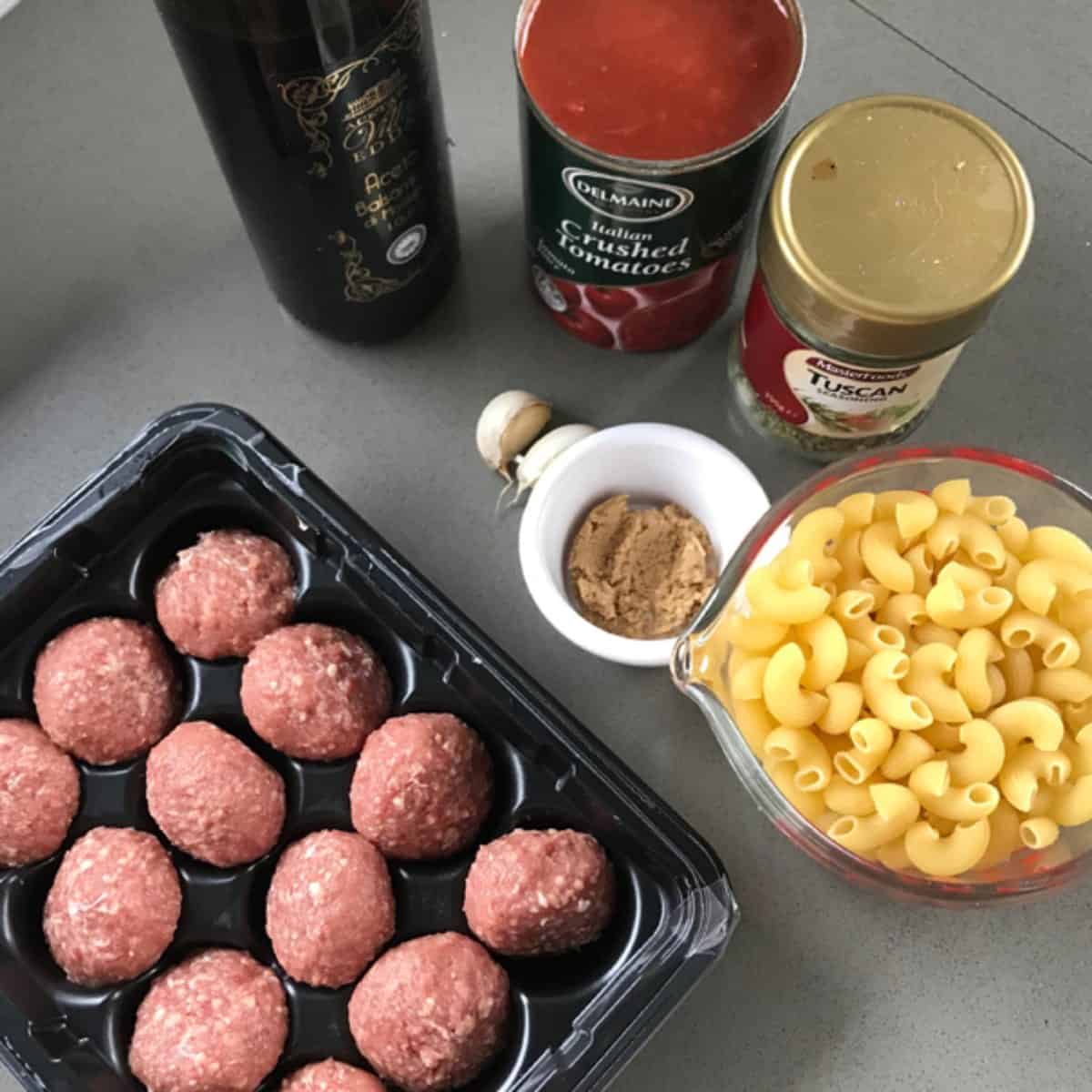 One Pot Baked Meatballs and Pasta Tray of raw meatballs, measuring cup of uncooked pasta, can of crushed tomatoes, jar of herbs, bottle of oil, garlic clove, and small bowl of brown spice set out for One Pot Baked Meatballs and Pasta on a gray countertop.