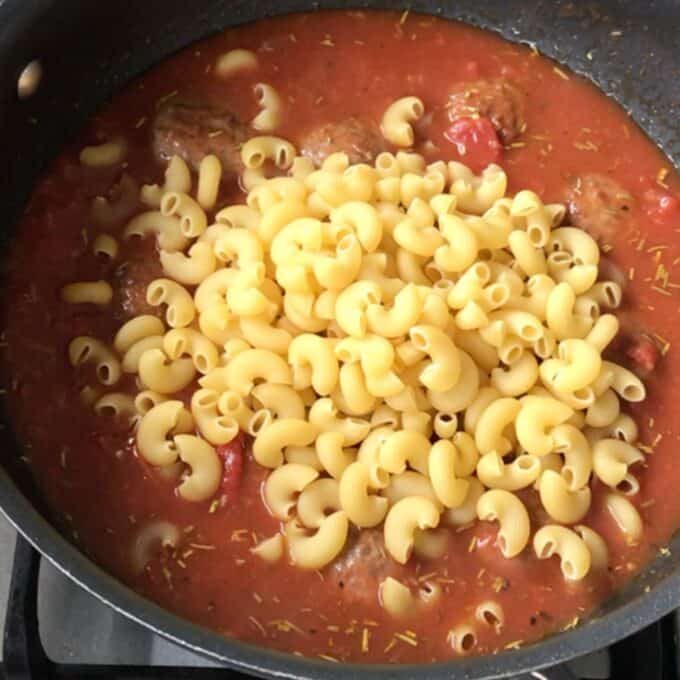 One Pot Baked Meatballs and Pasta A skillet on a stove contains tomato sauce with meatballs and uncooked elbow macaroni on top, ready to be mixed for an easy One Pot Baked Meatballs and Pasta dinner.