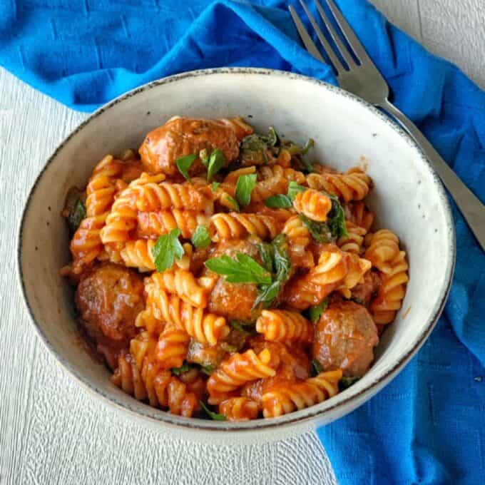 One Pot Baked Meatballs and Pasta A bowl of One Pot Baked Meatballs and Pasta in tomato sauce, garnished with fresh parsley, sits on a blue napkin with a fork beside it.