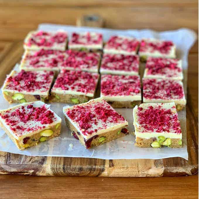 Cranberry and pistachio slice squares of white chocolate, studded with pistachios and cranberries, topped with a layer of crushed raspberries, are arranged on parchment paper atop a wooden cutting board.