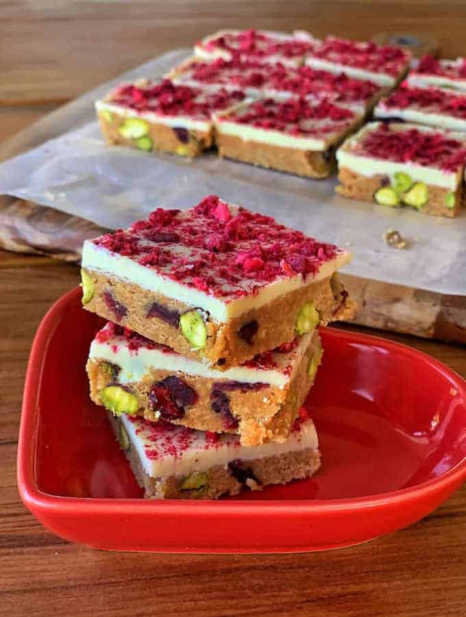 3 pieces of Cranberry and pistachio slice stacked on red plate with more pieces on a board in the background.