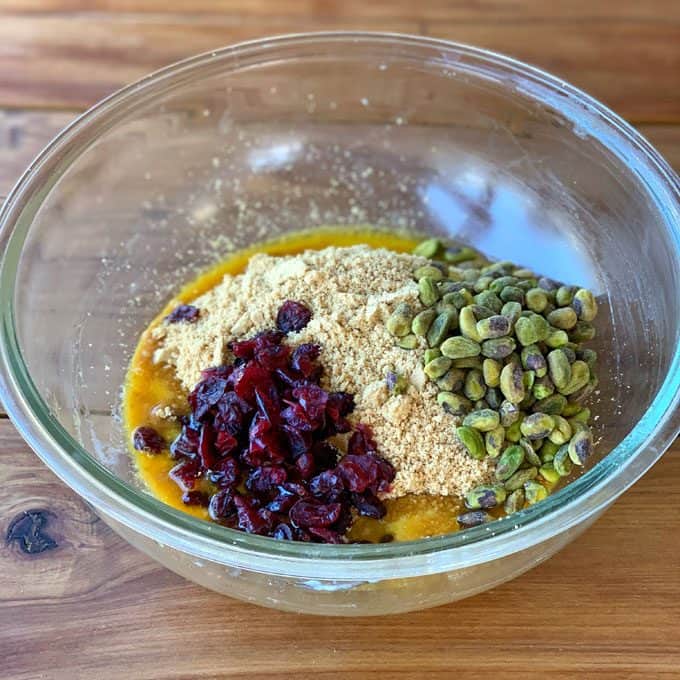 A glass bowl containing the ingredients for a cranberry and pistachio slice—melted butter, graham cracker crumbs, pistachios, and dried cranberries—sits on a wooden surface.