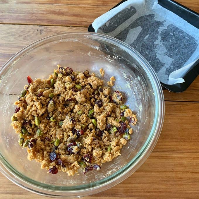 A glass bowl with granola bar mixture containing nuts and dried fruit, perfect for making cranberry and pistachio slice, sits on a wooden table next to a baking tray lined with parchment paper.