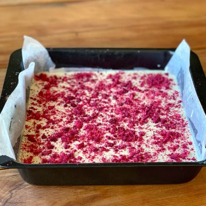 A square baking pan lined with parchment paper holds an unbaked cranberry and pistachio slice, topped with a layer of crushed red toppings, resting on a wooden surface.