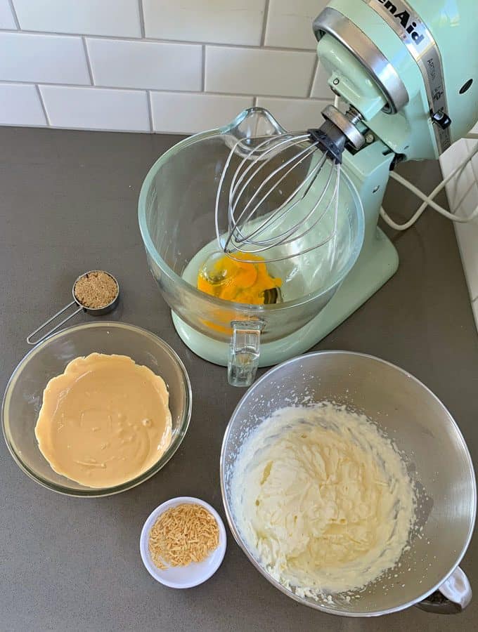 Caramilk Mousse A stand mixer with eggs and sugar in the bowl, surrounded by small bowls of brown sugar, sweetened condensed milk, whipped mixture, shredded coconut, and all the essentials for crafting a decadent Caramilk Mousse on the countertop.