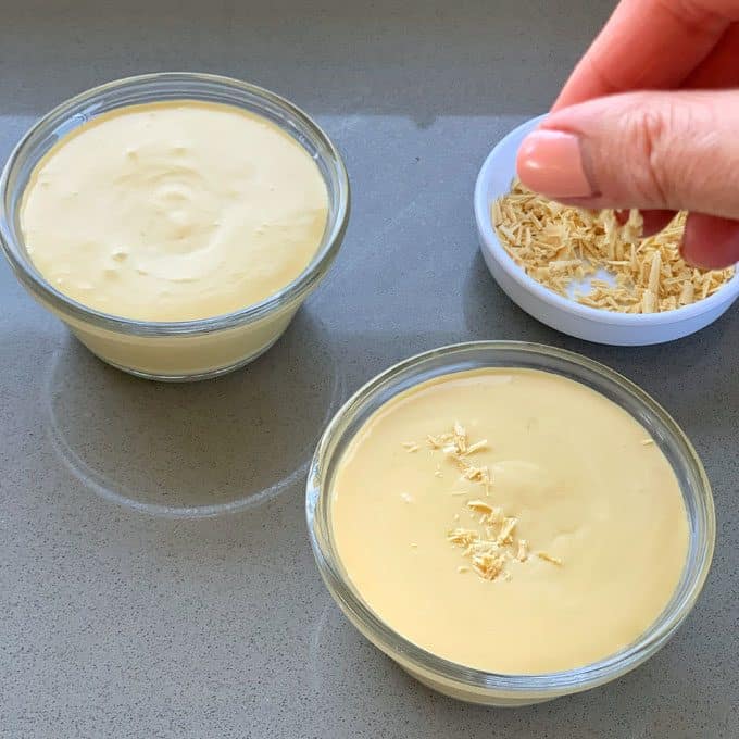 Caramilk Mousse A hand sprinkles shredded cheese onto creamy Caramilk Mousse in a glass bowl, with another bowl of mousse and a small dish of shredded cheese nearby.