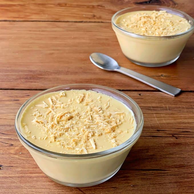 Caramilk Mousse Two glass bowls filled with creamy yellow Caramilk Mousse topped with grated white chocolate, placed on a wooden surface beside a metal spoon.