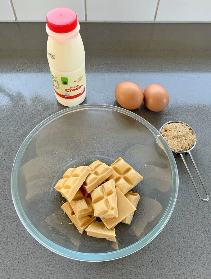 Caramilk Mousse A glass bowl with chunks of white chocolate, next to a measuring cup of brown sugar, two eggs, and a bottle of cream on a gray countertop—perfect ingredients for a decadent Caramilk Mousse.