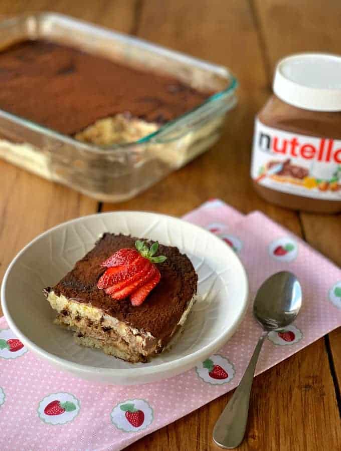 A piece of Nutellamisu in a white bowl with a spoon next to it on a wooden bench.