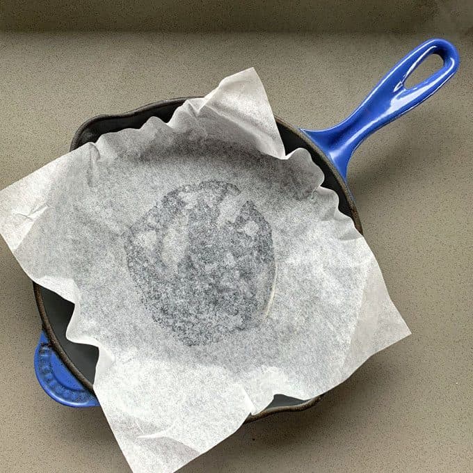 A parchment paper-lined cast iron skillet with a blue handle sits on a gray countertop, revealing the imprint of a freshly baked Chocolate and Raspberry Skillet Brownie.