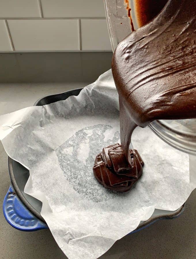 Brownie batter for a Chocolate and Raspberry Skillet Brownie is being poured from a bowl into a parchment-lined baking dish on a kitchen counter.