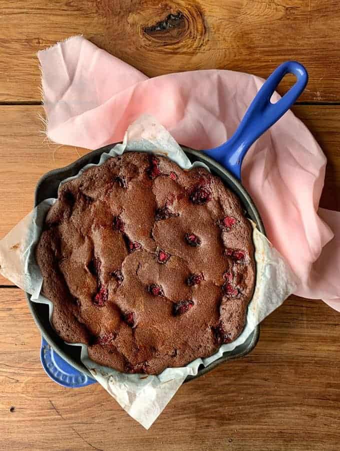 A Chocolate and Raspberry Skillet Brownie baked in a parchment-lined blue cast iron skillet, topped with fresh raspberries, sits on a wooden surface with a pink cloth nearby.
