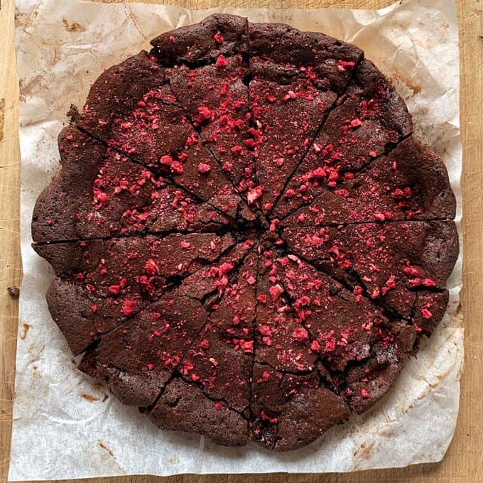 Chocolate and Raspberry Skillet Brownie A round, sliced Chocolate and Raspberry Skillet Brownie topped with crushed red berries sits on parchment paper atop a wooden surface.