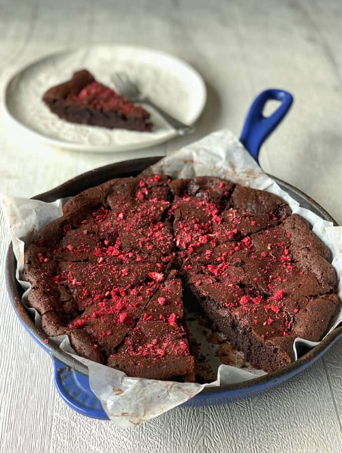 A round Chocolate and Raspberry Skillet Brownie with a slice removed, topped with crushed red berries, sits in a blue skillet lined with parchment; a single slice is on a plate in the background.