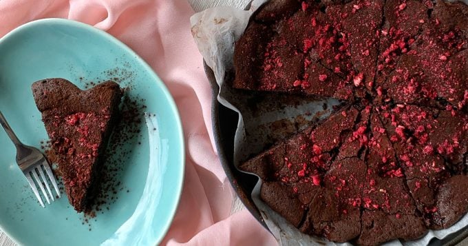 Chocolate and Raspberry Skillet Brownie A round Chocolate and Raspberry Skillet Brownie, partially sliced, sits on parchment paper with red berry pieces, and one slice served on a blue plate beside a fork.