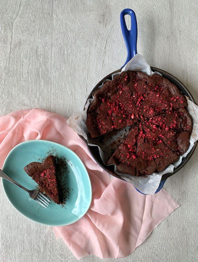 Chocolate and Raspberry Skillet Brownie A Chocolate and Raspberry Skillet Brownie sits in a blue skillet, sliced with one piece on a blue plate nearby; both are topped with red sprinkles and set on a light pink cloth.