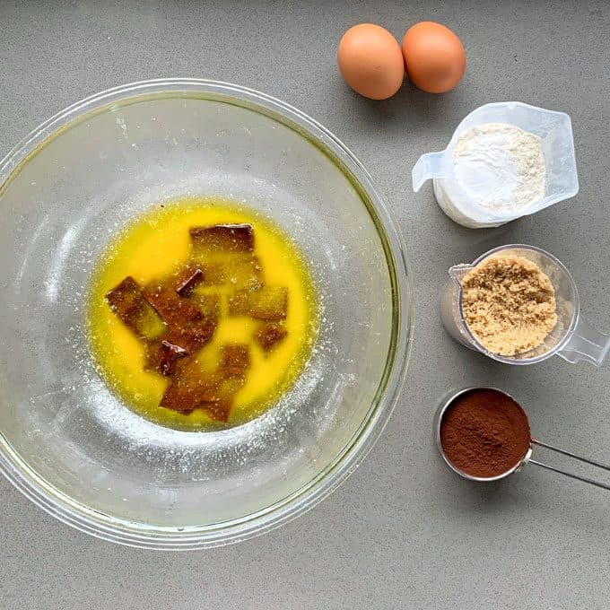 A glass bowl with melted butter and brown sugar sits beside two eggs, a small container of flour, brown sugar, and a measuring cup of cocoa powder—ingredients ready to create a delicious Chocolate and Raspberry Skillet Brownie.