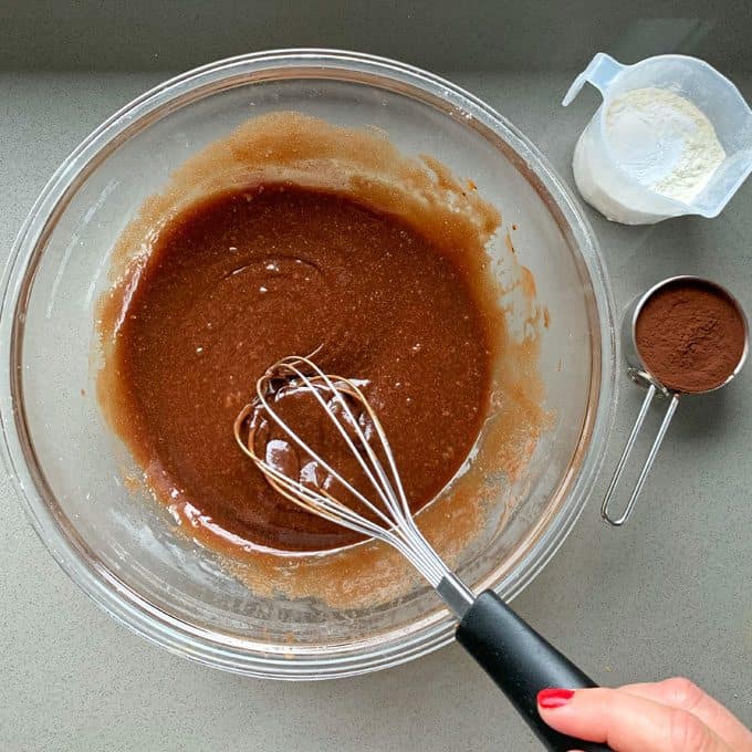 Chocolate and Raspberry Skillet Brownie A hand holding a whisk mixes chocolate batter in a glass bowl, with a measuring cup of flour and cocoa powder nearby—perfect ingredients for baking a rich Chocolate and Raspberry Skillet Brownie.