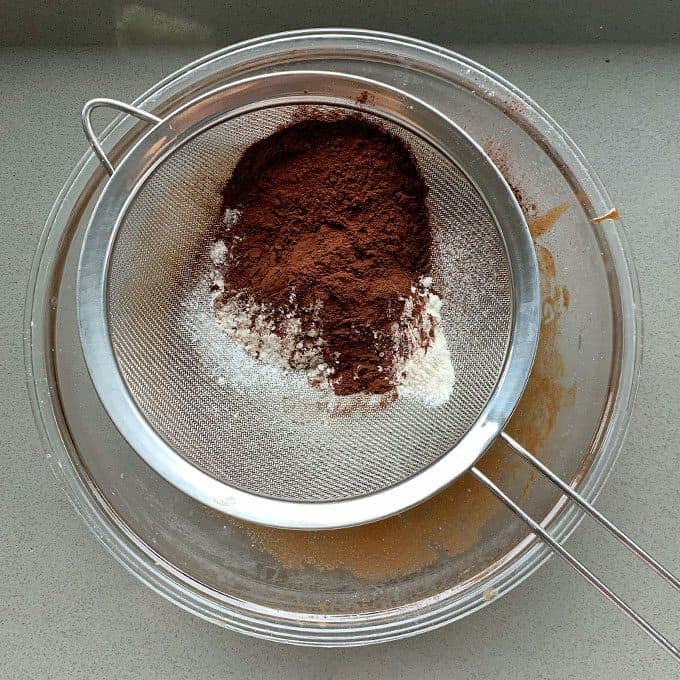 A mesh sieve containing cocoa powder and flour is placed over a glass mixing bowl, ready to create the base for a delicious Chocolate and Raspberry Skillet Brownie.