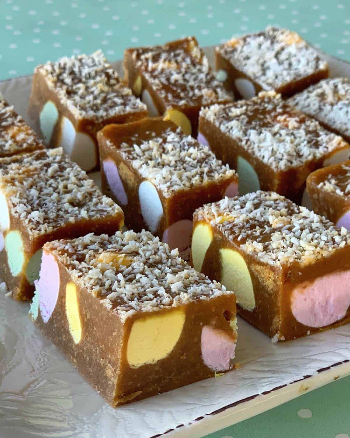 Squares of Lolly Slice with colored marshmallows and a coconut topping are arranged on a white plate.