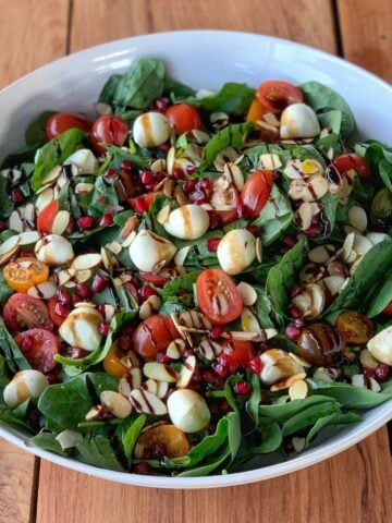A large white bowl filled with Christmas green salad-spinach, cherry tomatoes, mozzarella balls, sliced almonds, pomegranate seeds, and balsamic glaze-sits on a wooden table.