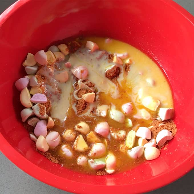 Lolly Slice A red mixing bowl containing the ingredients for lolly slice: melted butter, sweetened condensed milk, crushed biscuits, and colorful mini marshmallows.
