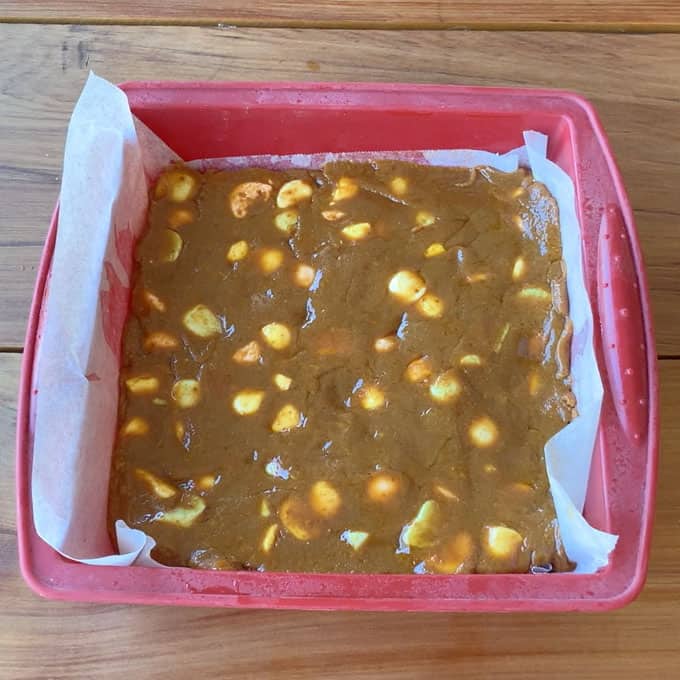 Lolly Slice A red square baking dish lined with parchment paper is filled with unbaked lolly slice batter, visible chunks peeking through, and sits on a wooden surface.