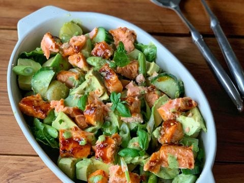 A white oval dish filled with a grilled salmon and potato salad, featuring avocado, cucumber, and leafy greens, set on a wooden table with two serving utensils beside it.