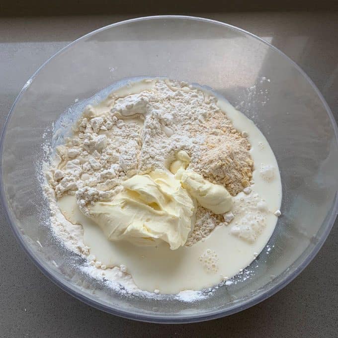 A clear bowl containing flour, milk, butter, and dry ingredients for a delicious sausage and herb loaf sits ready to be mixed on a light-colored countertop.