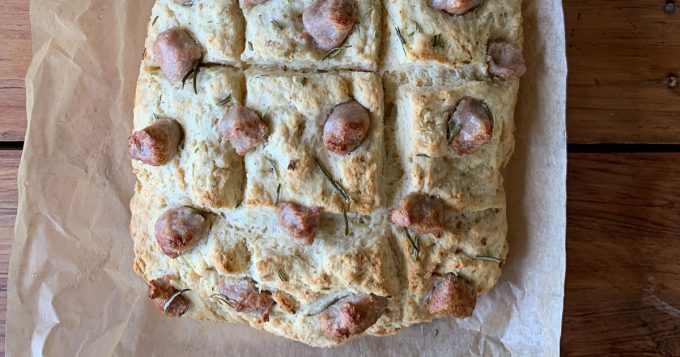 sausage and herb loaf A sausage and herb loaf of focaccia bread on parchment paper, topped with sprigs of rosemary and small browned sausage pieces, shown from above.