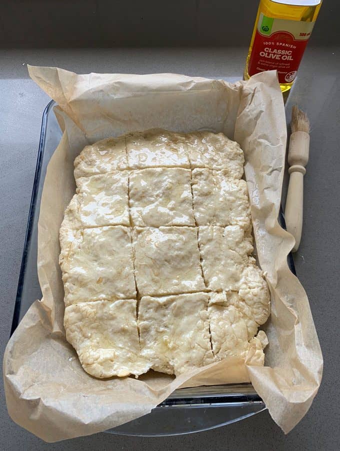 A parchment-lined baking dish with unbaked sausage and herb loaf dough scored into squares, a pastry brush, and a bottle of olive oil on a gray countertop.