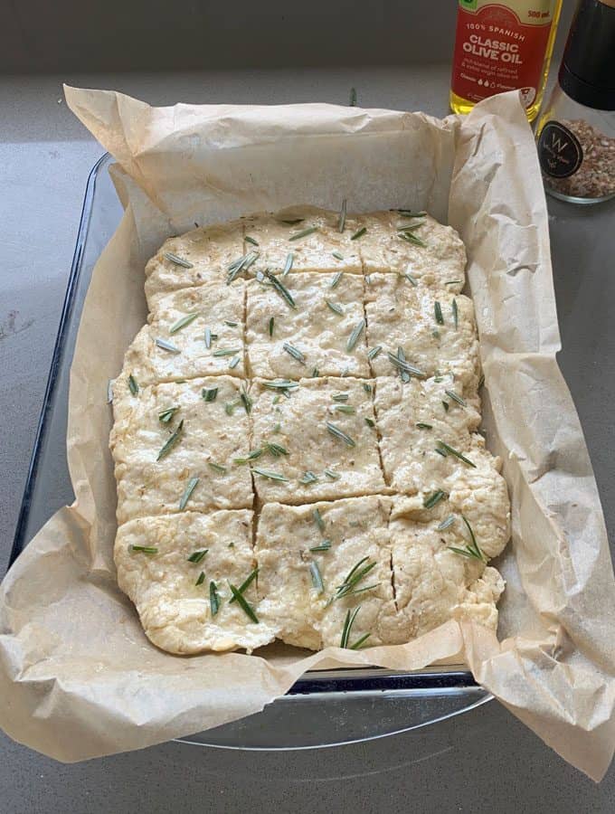 sausage and herb loaf Unbaked focaccia dough in a parchment-lined baking dish, topped with rosemary and scored into squares, sits on a gray countertop next to a bottle of olive oil and a pepper grinder—perfect inspiration for any sausage and herb loaf.