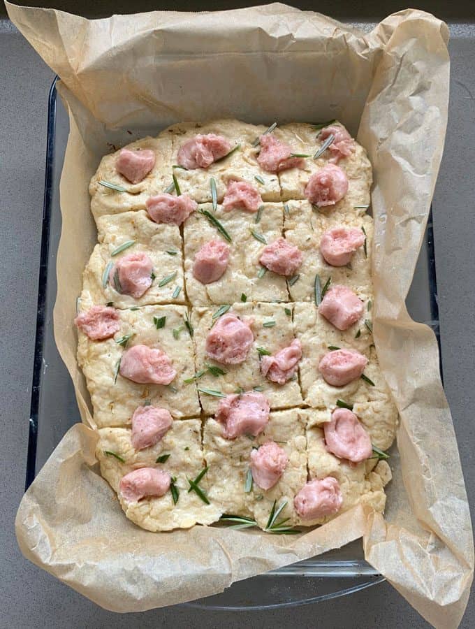 A baking dish lined with parchment paper holds unbaked focaccia dough, pre-sliced into squares and topped with pink dollops and rosemary, echoing the savory appeal of a sausage and herb loaf.