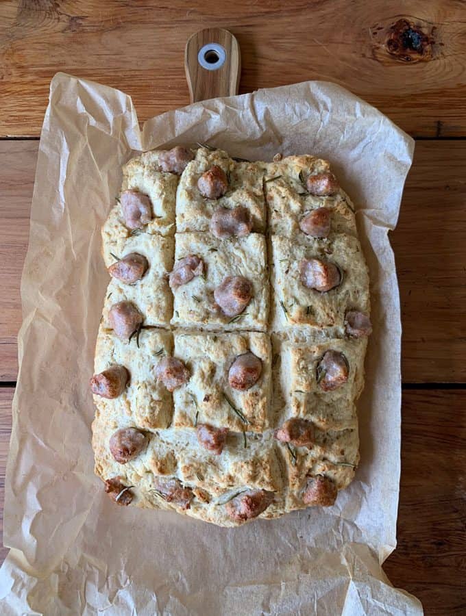 sausage and herb loaf A rectangular, rustic sausage and herb loaf focaccia on parchment paper rests on a wooden cutting board, with visible dents and a golden-brown top.