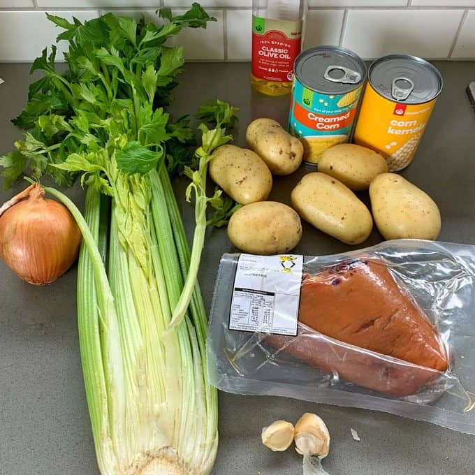 Corn and potato chowder with bacon hock A bundle of celery, an onion, several potatoes, two cans of corn—perfect for making corn and potato chowder—plus a bottle of olive oil, garlic cloves, packaged smoked fish, and bacon hock on a kitchen counter.