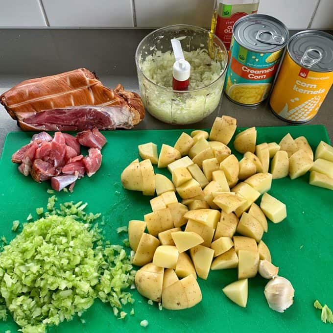 Chopped potatoes, celery, onion, and ham are on a cutting board with garlic and canned corn, creamed corn, and a bacon hock nearby—perfect ingredients for a hearty corn and potato chowder.