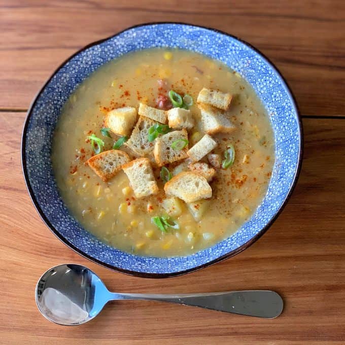 Corn and potato chowder with bacon hock A bowl of creamy corn and potato chowder with bacon hock, topped with croutons, chopped green onions, and a sprinkle of paprika, served on a wooden table with a spoon beside it.