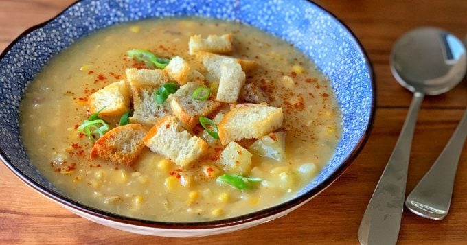 A bowl of thick corn and potato chowder with bacon hock, topped with croutons and chopped green onions, sits on a wooden table beside a spoon and fork.
