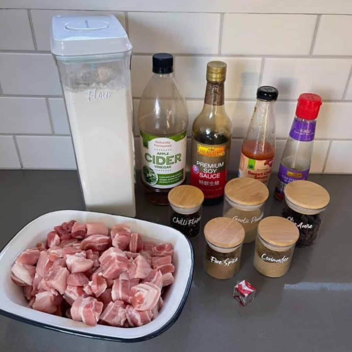 Raw cubed pork, flour, sauces, spices, and a stock cube are arranged on a kitchen counter in front of a tiled backsplash—perfect for preparing Sticky Chinese Pork Belly.
