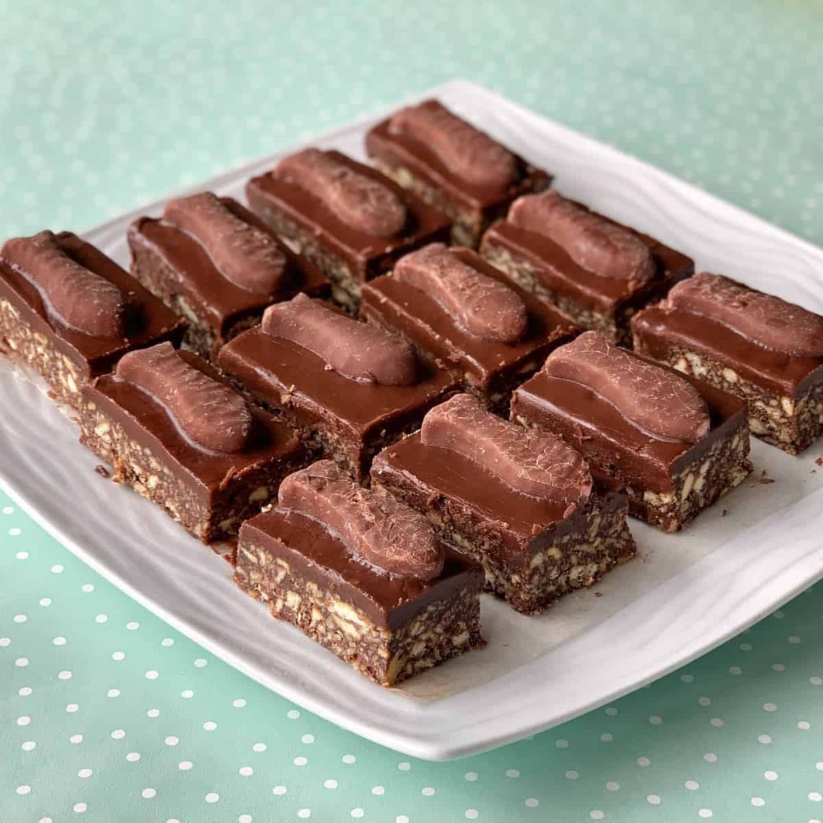 A plate of rectangular chocolate fish slice biscuits, topped with chocolate bars and arranged in rows on a white plate, sits against a light green polka dot background.