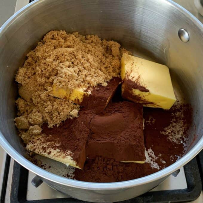A saucepan on a stove contains brown sugar, cocoa powder, and large chunks of butter, ready to be mixed or melted together for a delicious chocolate fish slice.