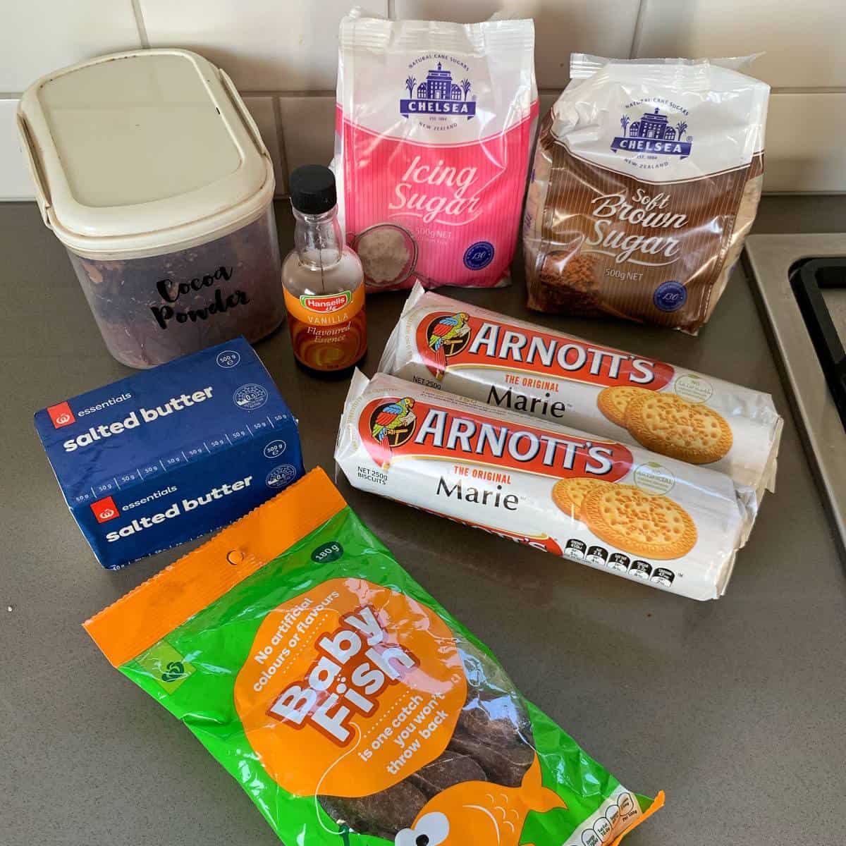 Chocolate Fish Slice Various baking ingredients on a kitchen counter, including cocoa powder, icing sugar, brown sugar, vanilla extract, salted butter, Marie biscuits, and a packet of Baby Fish snacks—perfect for whipping up a delicious chocolate fish slice.
