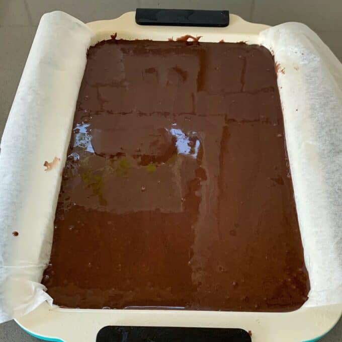 A rectangular baking tray lined with parchment paper is filled with unbaked chocolate brownie batter, ready to be smoothed with a chocolate fish slice.