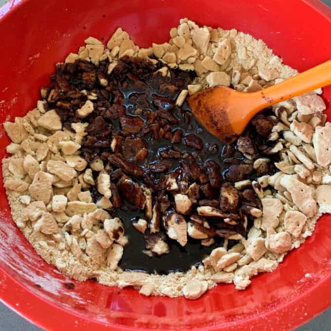 Chocolate Fish Slice A red mixing bowl filled with broken biscuit pieces and a dark chocolate fish slice mixture, being stirred with an orange spatula.